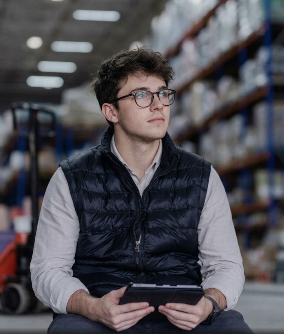 A new salesperson in glasses and a gilet sits in a warehouse aisle with a tablet, looking up at the racking
